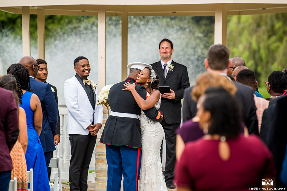  bride in a white formfitting gown with a plunging neckline and lace detailing and the groom in a white shawl lapel tuxedo with a black shirt and gold bow tie, giving away the bride at the ceremony