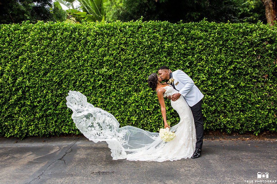  bride in a white formfitting gown with a plunging neckline and lace detailing and the groom in a white shawl lapel tuxedo with a black shirt and gold bow tie, kissing by ivy wall