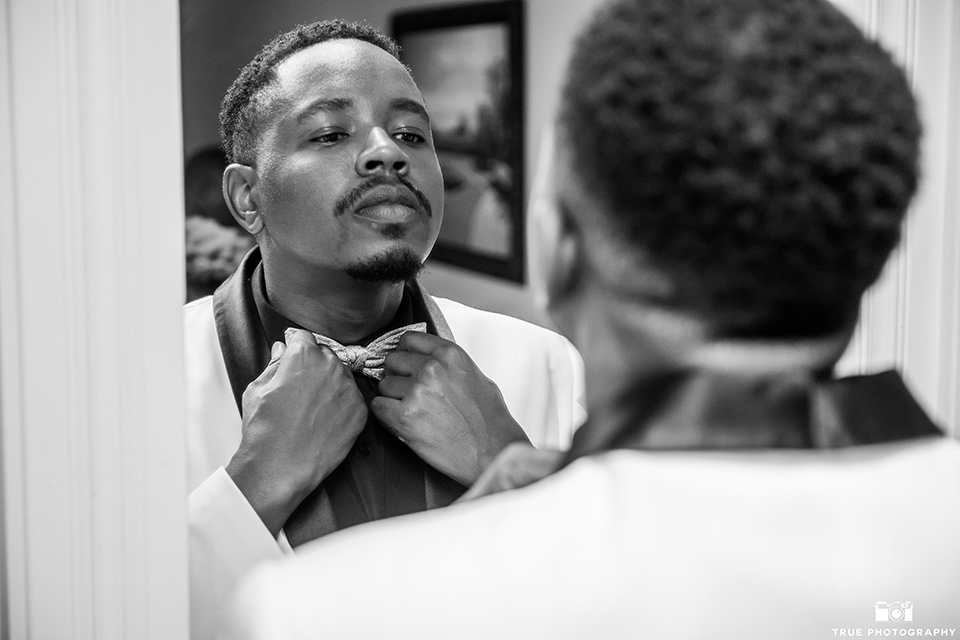  the groom in a white shawl lapel tuxedo with a black shirt and gold bow tie putting on his bow tie