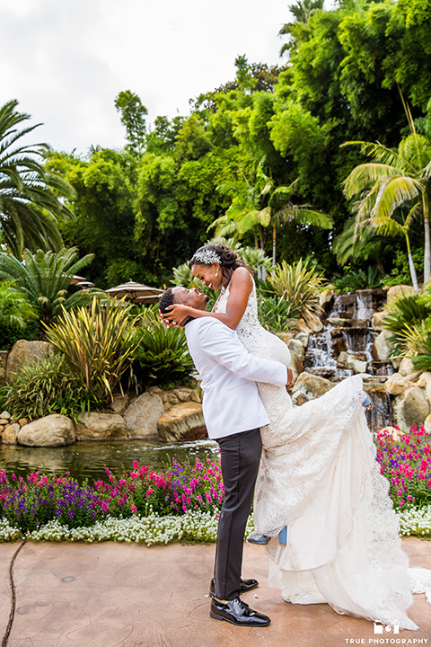  bride in a white formfitting gown with a plunging neckline and lace detailing and the groom in a white lace tuxedo with a black shirt and gold bow tie, groom lifting up the bride