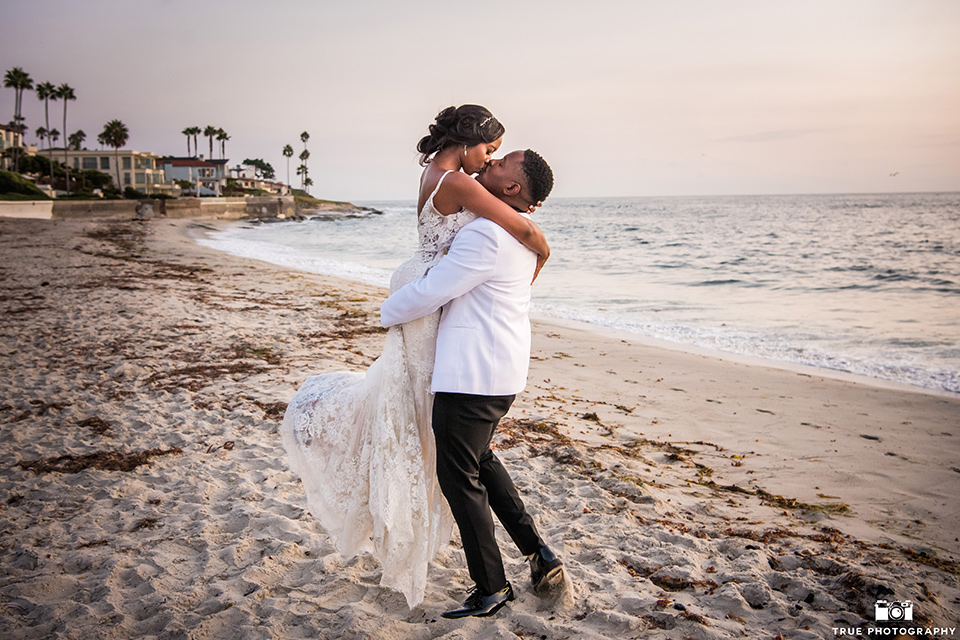  bride in a white formfitting gown with a plunging neckline and lace detailing and the groom in a white shawl lapel tuxedo with a black shirt and gold bow tie, kissing on the beach