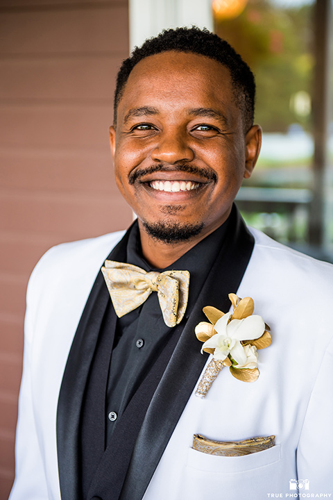  the groom in a white lace tuxedo with a black shirt and gold bow tie, smiling at the camera