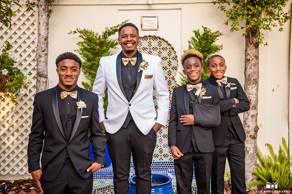  the groom in a white shawl lapel tuxedo with a black shirt and gold bow tie and groomsmen in all black tuxedos with gold bow ties