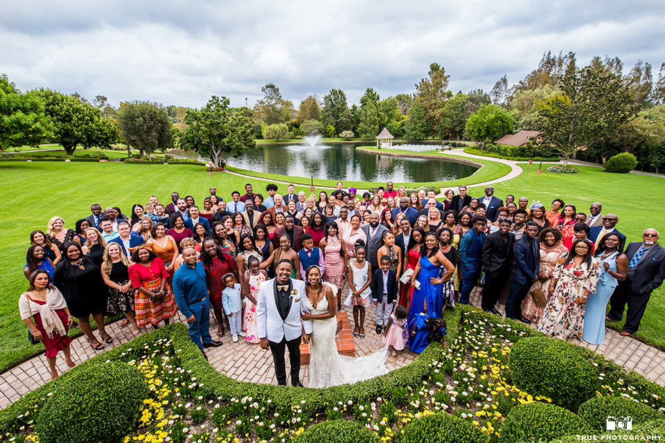  bride in a white formfitting gown with a plunging neckline and lace detailing and the groom in a white shawl lapel tuxedo with a black shirt and gold bow tie, with their entire wedding guests in one picture