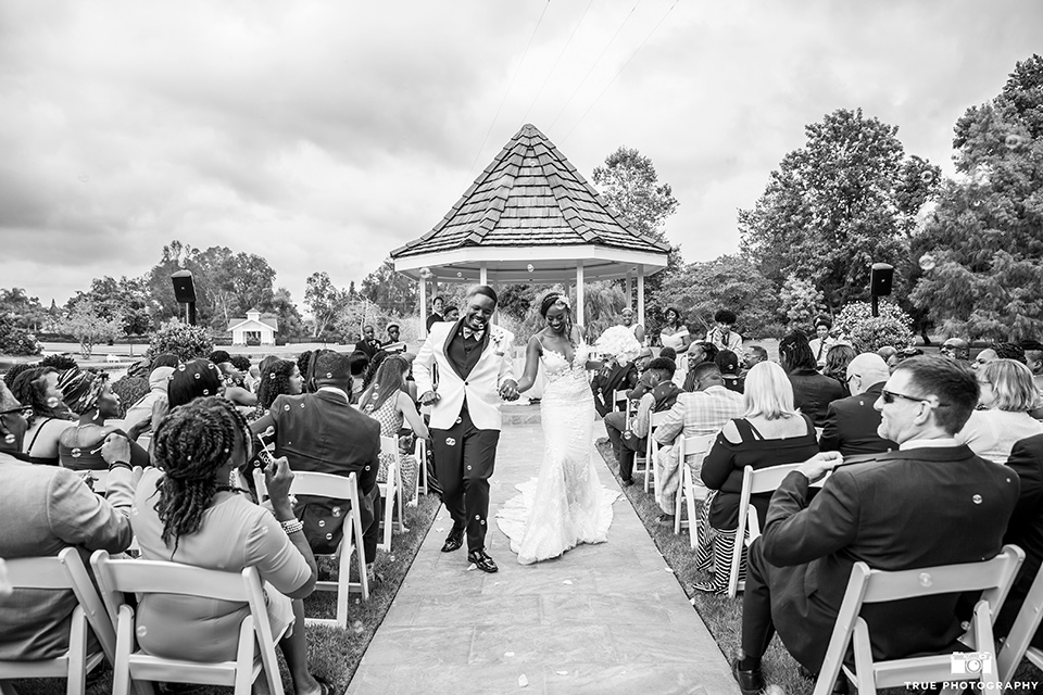  bride in a white formfitting gown with a plunging neckline and lace detailing and the groom in a white shawl lapel tuxedo with a black shirt and gold bow tie, walking down the aisle