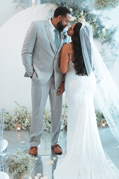  bride in a formfitting satin gown and a cathedral length veil and the groom in a light grey peak lapel suit with a teal blue bow tie 