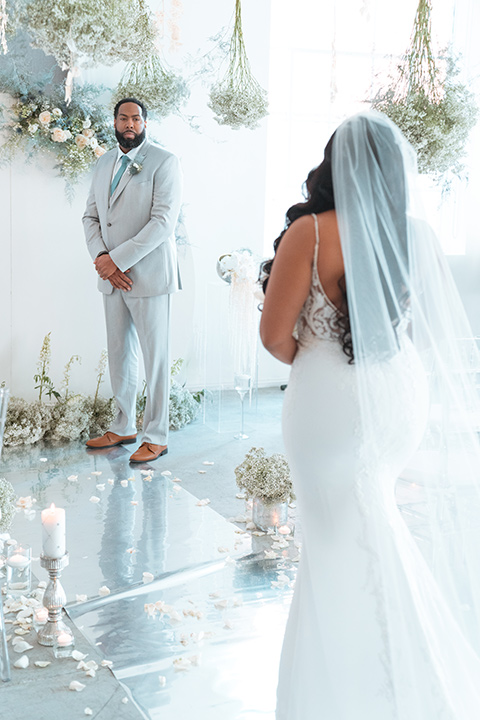  bride in a formfitting satin gown and a cathedral length veil and the groom in a light grey peak lapel suit with a teal blue bow tie 