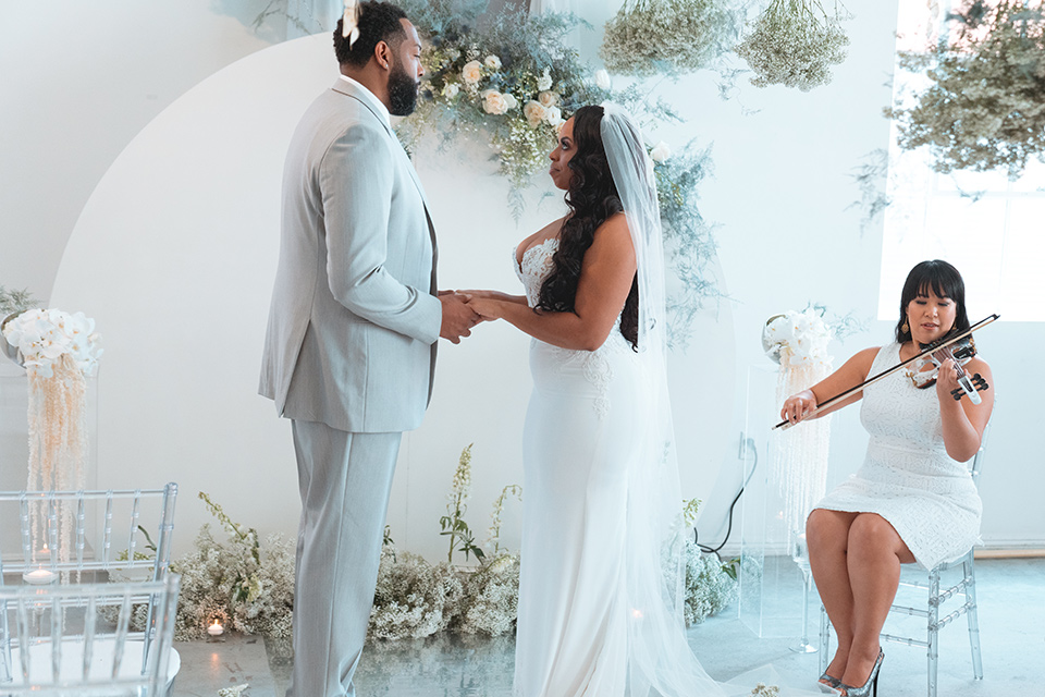  bride in a formfitting satin gown and a cathedral length veil and the groom in a light grey peak lapel suit with a teal blue bow tie 