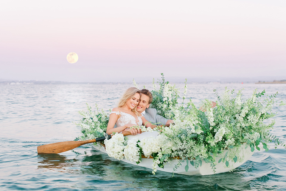  bride in a white formfitting lace gown with an off the shoulder detailing and her hair in loose waves, the groom in a light grey suit with a white long tie sitting in a boat together