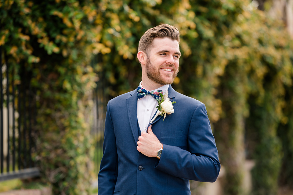  groom in a dark blue suit with a blue bow tie and the bride in a lace gown with a high neckline, and the groomsmen in a blue suit with green bow ties and bridesmaids in green/teal gowns
