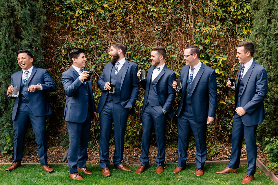  groom in a dark blue suit with a blue bow tie and the bride in a lace gown with a high neckline, and the groomsmen in a blue suit with green bow ties and bridesmaids in green/teal gowns
