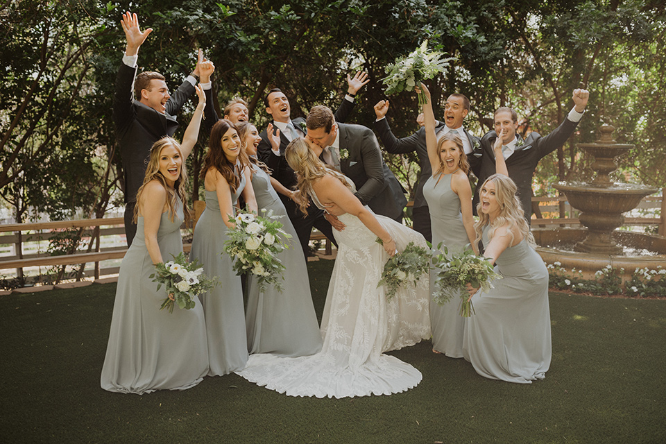  =” the bride in a flowing white gown with a high lace neckline and a long cathedral veil, the groom in an asphalt grey suit by Michael Kors with a light blue patterned long tie, the bridesmaids in dusty blue long gowns and the groomsmen in dark grey suits with floral long ties