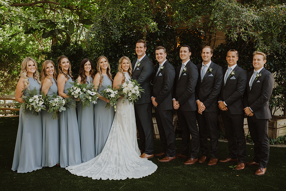 =” the bride in a flowing white gown with a high lace neckline and a long cathedral veil, the groom in an asphalt grey suit by Michael Kors with a light blue patterned long tie, the bridesmaids in dusty blue long gowns and the groomsmen in dark grey suits with floral long ties