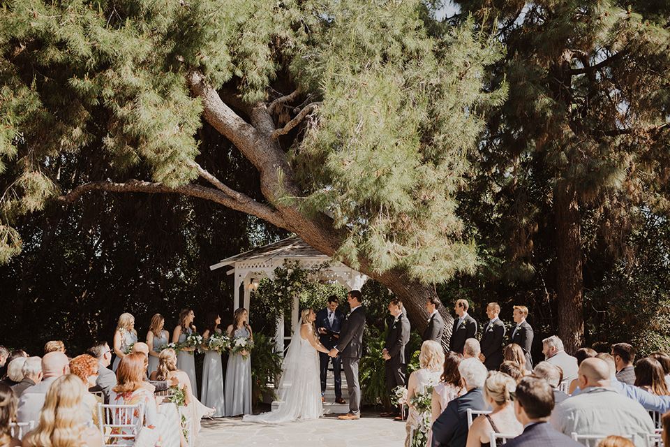  =” the bride in a flowing white gown with a high lace neckline and a long cathedral veil, the groom in an asphalt grey suit by Michael Kors with a light blue patterned long tie, the bridesmaids in dusty blue long gowns and the groomsmen in dark grey suits with floral long ties at the ceremony