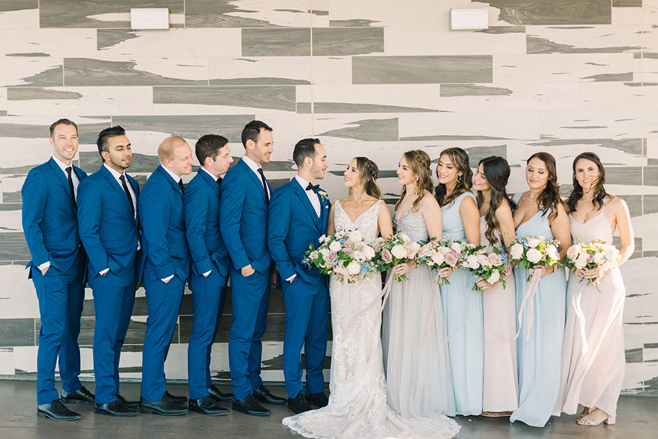  bride in an ivory lace gown with a modern geometric pattern and a v neckline, the groom wore a cobalt blue suit with a black bowtie, the bridesmaids wore different color dresses in pastel tones and the groomsmen in cobalt suits with black long ties