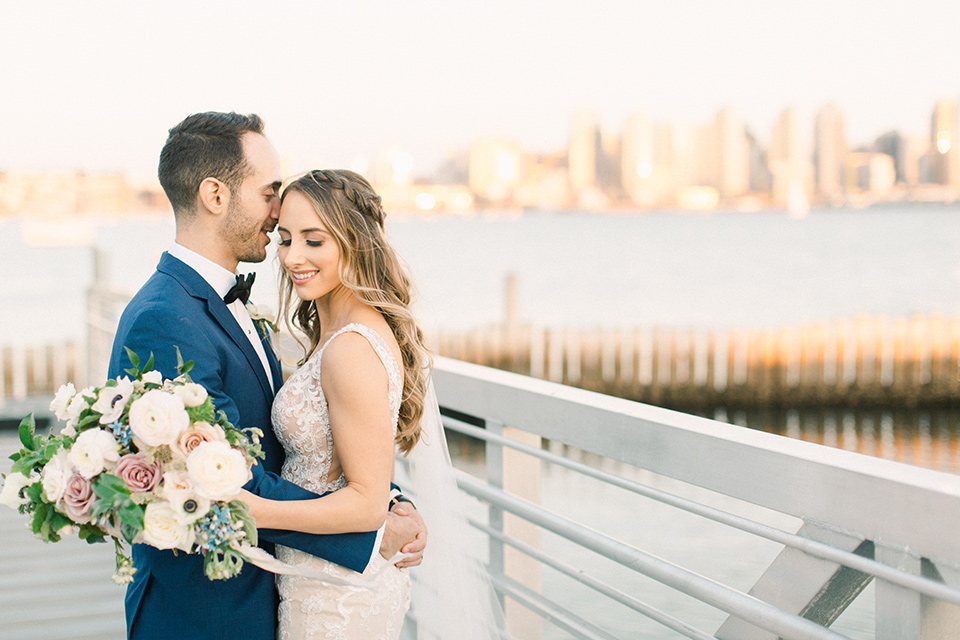  bride in an ivory lace gown with a modern geometric pattern and a v neckline, the groom wore a cobalt blue suit with a black bowtie with skyline and ocean behind them 