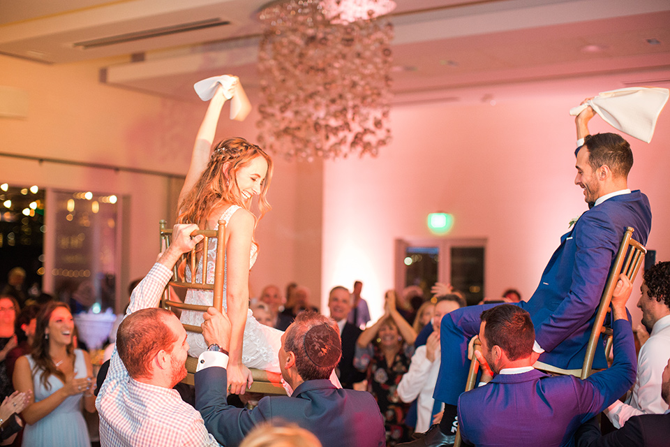  bride in an ivory lace gown with a modern geometric pattern and a v neckline, the groom wore a cobalt blue suit with a black bowtie on chairs being held by wedding guests