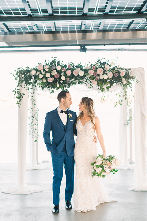  bride in an ivory lace gown with a modern geometric pattern and a v neckline, the groom wore a cobalt blue suit with a black bowtie walking by ceremony space