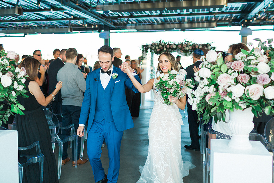  bride in an ivory lace gown with a modern geometric pattern and a v neckline, the groom wore a cobalt blue suit with a black bowtie walking out of ceremony