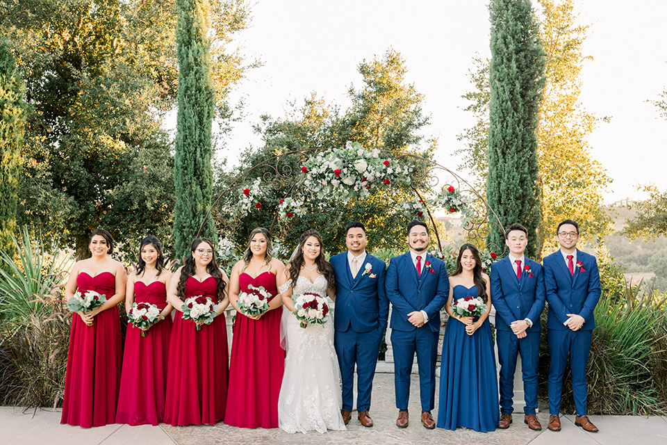 bride in a Spanish style lace gown with off the shoulder detailing, the groom in a cobalt suit with a pink long tie, the groomsmen in cobalt suits with red long ties, and bridesmaids in red dresses
