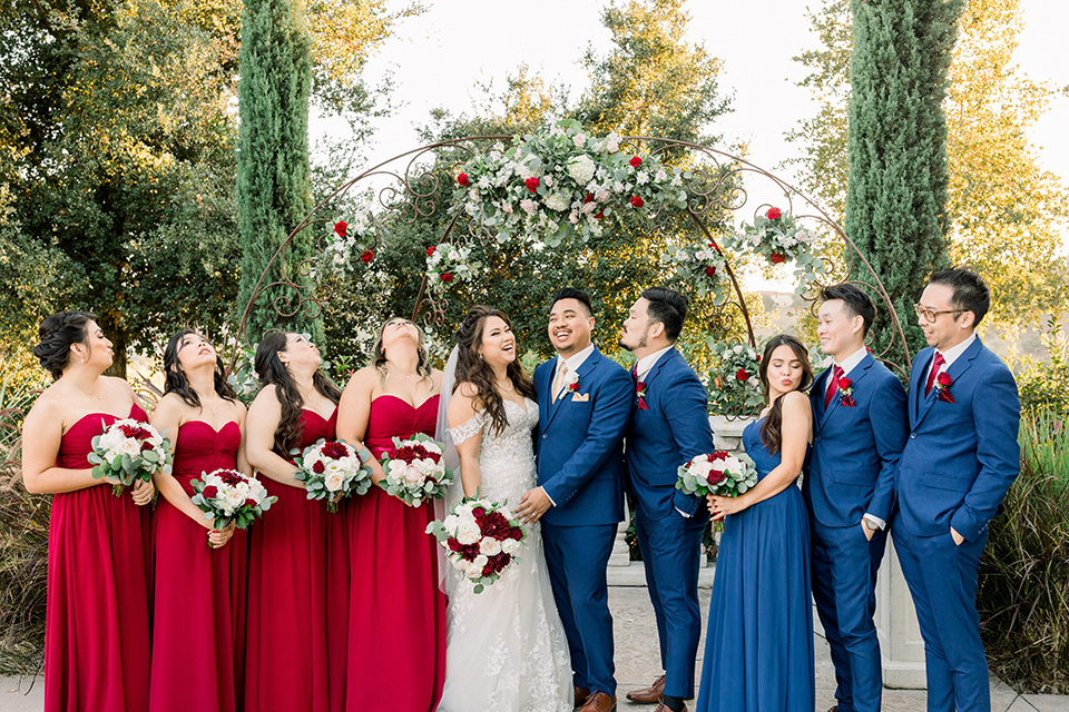 bride in a Spanish style lace gown with off the shoulder detailing, the groom in a cobalt suit with a pink long tie, the groomsmen in cobalt suits with red long ties, and bridesmaids in red dresses