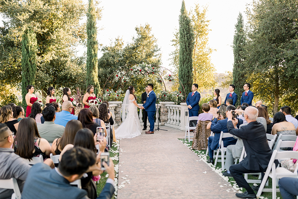  bride in a Spanish style lace gown with off the shoulder detailing, the groom in a cobalt suit with a pink long tie, the groomsmen in cobalt suits with red long ties, and bridesmaids in red dresses at ceremony