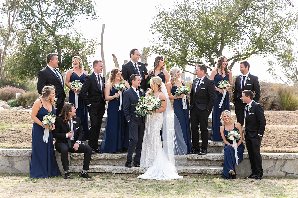  bride in a white lace gown and the groom in a navy shawl lapel tuxedo and blow tie, bridesmaids in navy gowns and groomsmen in black tuxedos