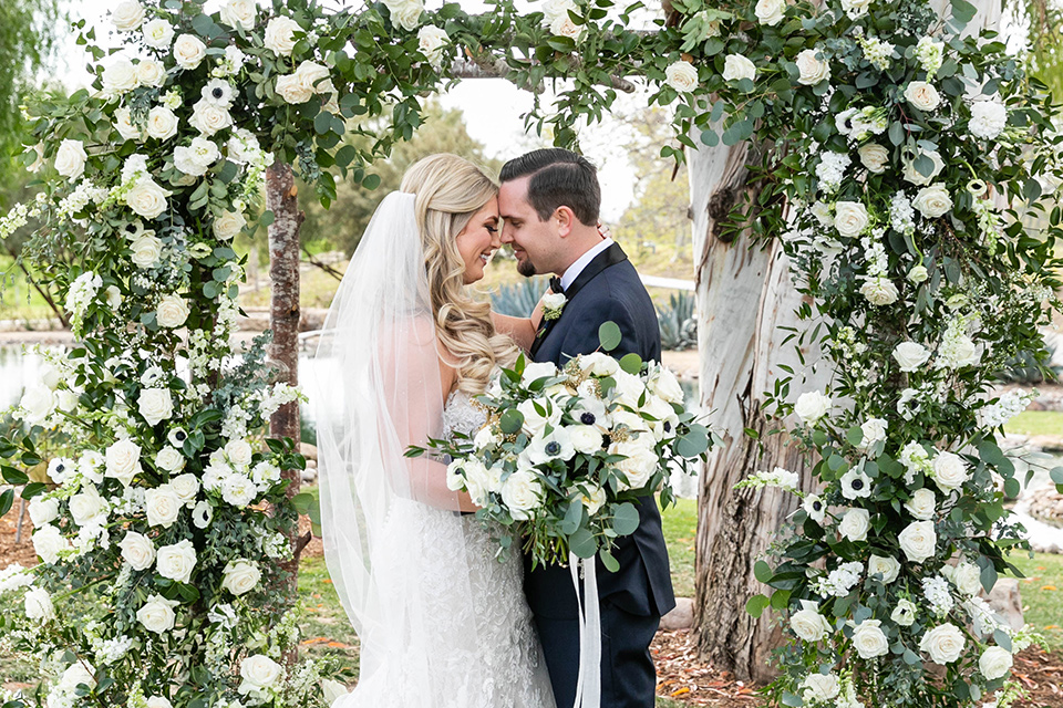  bride in a white lace gown and the groom in a navy shawl lapel tuxedo and blow tie, by ceremony arch