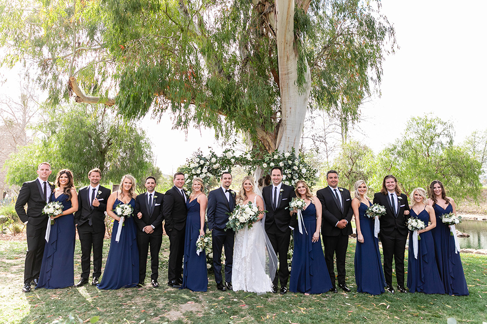  bride in a white lace gown and the groom in a navy shawl lapel tuxedo and blow tie, bridesmaids in navy gowns and groomsmen in black tuxedos