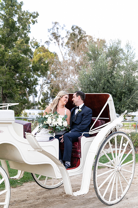  bride in a lace gown and long veil, groom in a navy shawl lapel tuxedo with a black bow tie in horse drawn carriage 