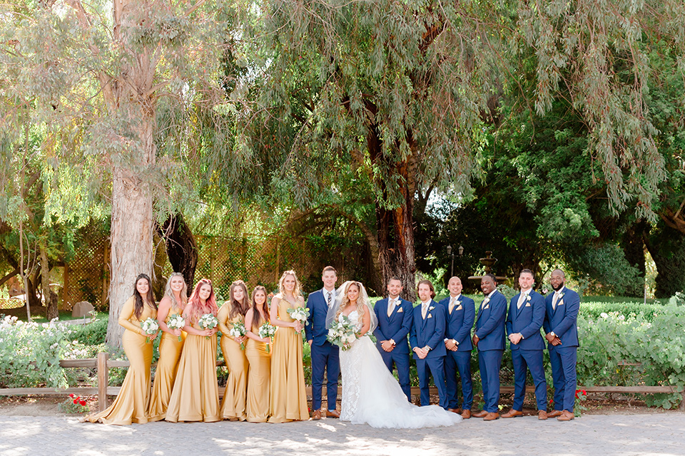  bride in a white lace gown with a long train and veil, groom in a cobalt blue suit with a blue floral long tie, the bridesmaids in a gold ballgowns and groomsmen in cobalt blue suit with blue floral bow ties 