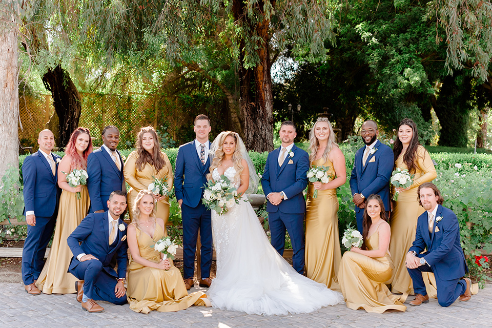  bride in a white lace gown with a long train and veil, groom in a cobalt blue suit with a blue floral long tie, the bridesmaids in a gold ballgowns and groomsmen in cobalt blue suit with blue floral bow ties 
