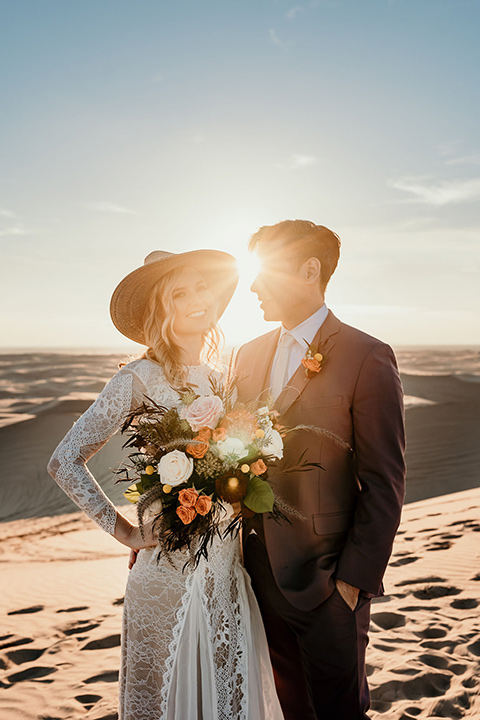  bride in a lace gown with long sleeves and a wide brimmed hat and the groom in a rose pink suit with a white long tie