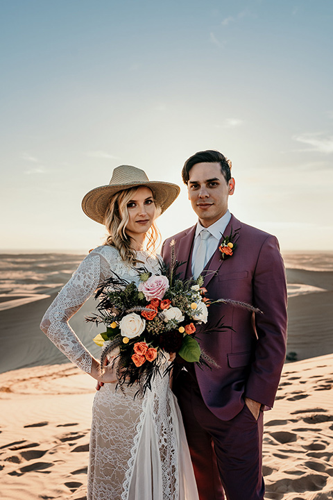  bride in a lace gown with long sleeves and a wide brimmed hat and the groom in a rose pink suit with a white long tie 