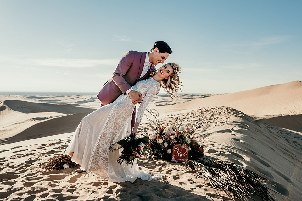  bride in a lace gown with long sleeves and a wide brimmed hat and the groom in a rose pink suit with a white long tie 