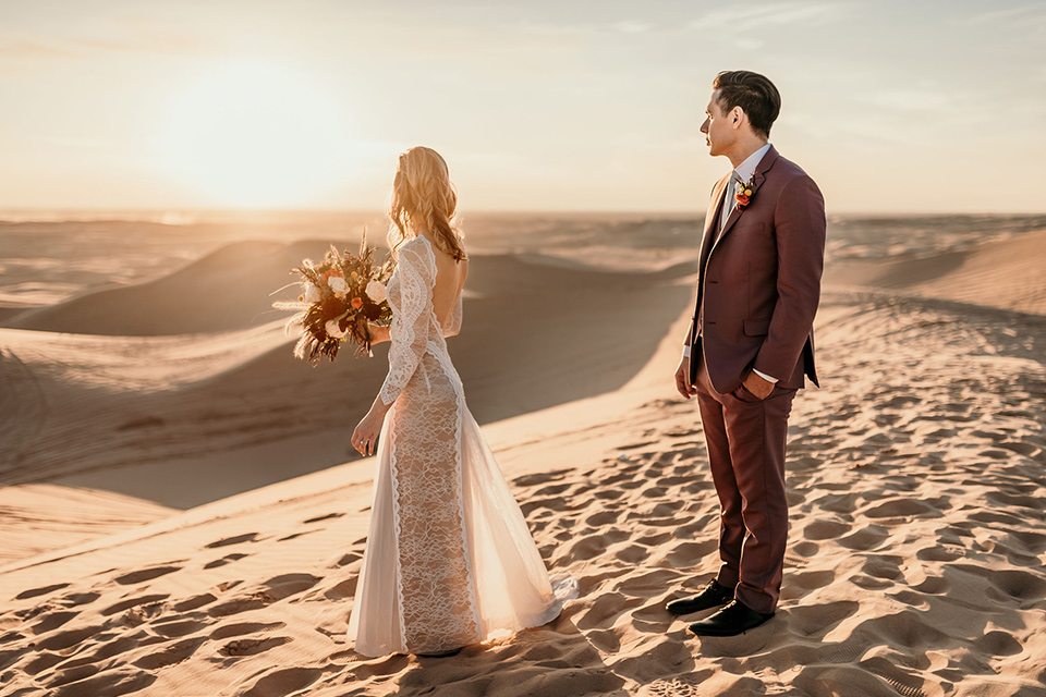  bride in a lace gown with long sleeves and a wide brimmed hat and the groom in a rose pink suit with a white long tie 