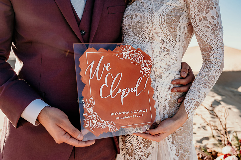  bride in a lace gown with long sleeves and a wide brimmed hat and the groom in a rose pink suit with a white long tie 