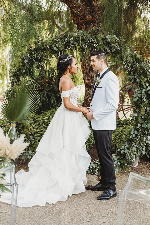  bride in a lace ballgown with off the shoulder neckline and groom in a white with a black shawl lapel tuxedo at ceremony