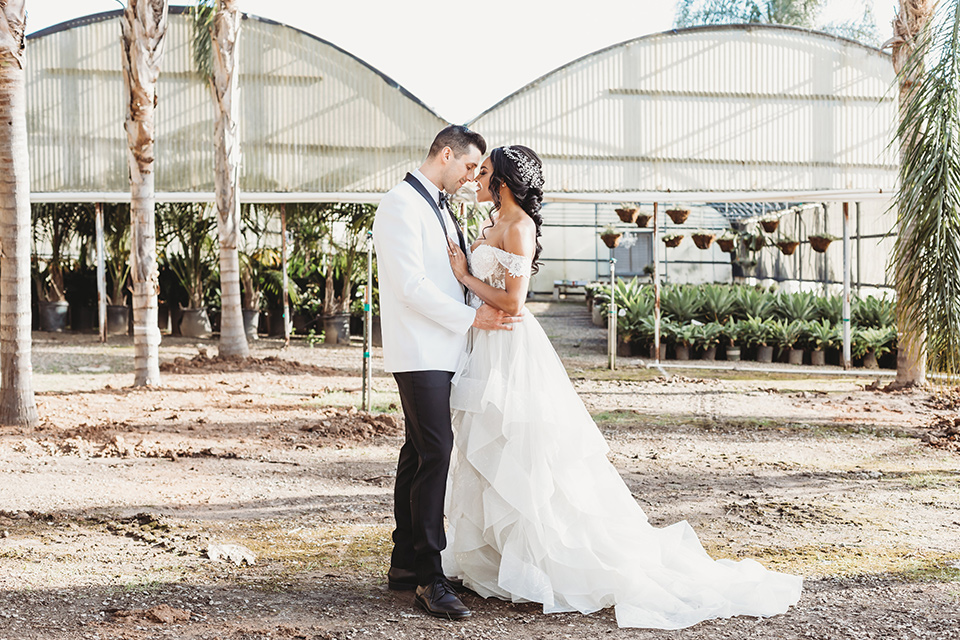  bride in a lace ballgown with off the shoulder neckline and groom in a white with a black shawl lapel tuxedo embracing outside the venue