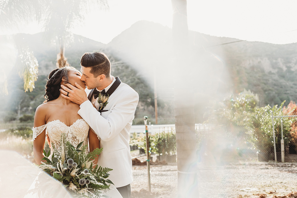  bride in a lace ballgown with off the shoulder neckline and groom in a white with a black shawl lapel tuxedo embracing outside the venue