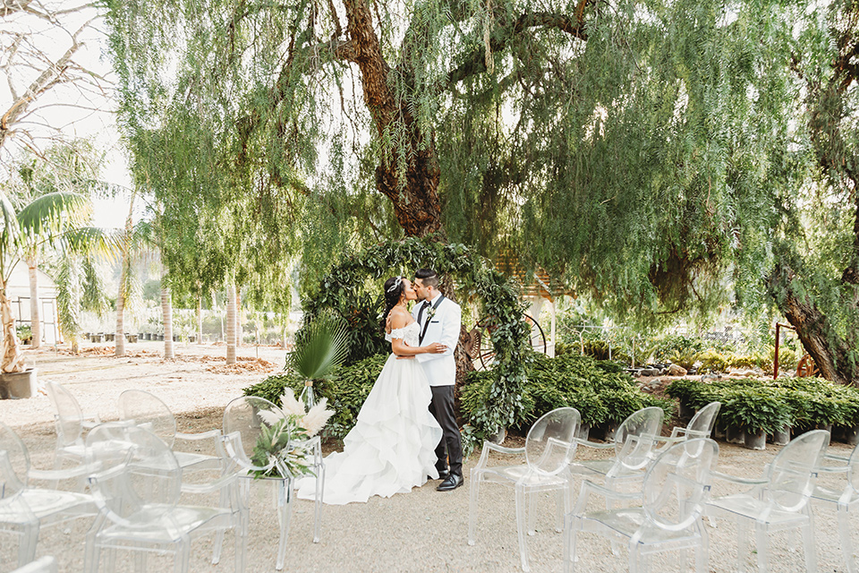  bride in a lace ballgown with off the shoulder neckline and groom in a white with a black shawl lapel tuxedo embracing at ceremony