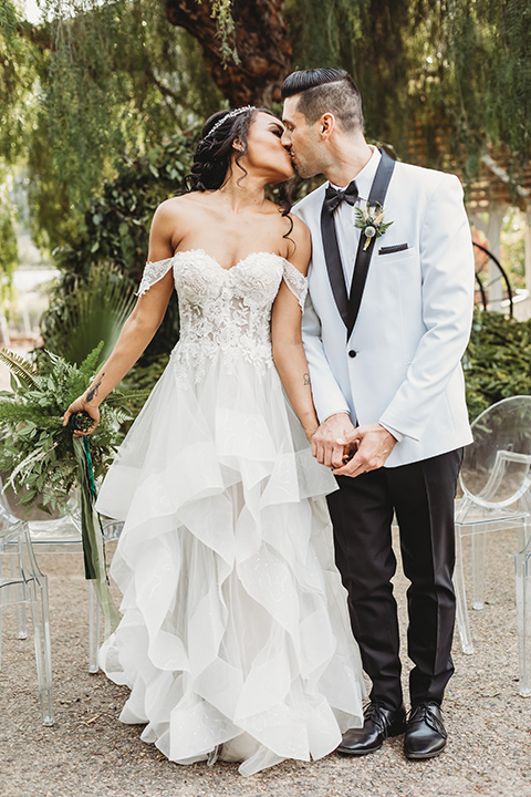  bride in a lace ballgown with off the shoulder neckline and groom in a white with a black shawl lapel tuxedo at ceremony