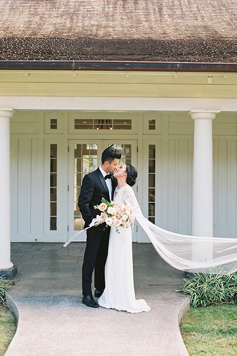  bride in a white formfitting gown with a long ethereal cape and her hair in a low bun and the groom in a black notch lapel tuxedo with bow tie outside pf venue