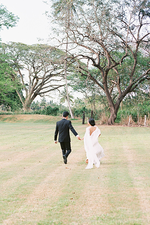  bride in a white formfitting gown with a long ethereal cape and her hair in a low bun and the groom in a black notch lapel tuxedo with bow tie outside pf venue