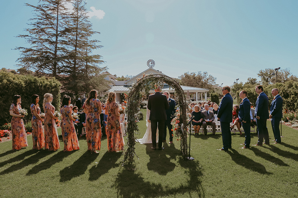  bride in a lace bohemian gown with tulip sleeves and a braided crown, the groom in a cobalt blue suit for purchase and the groomsmen in rental cobalt suits and the bridesmaids in pink floral gowns 
