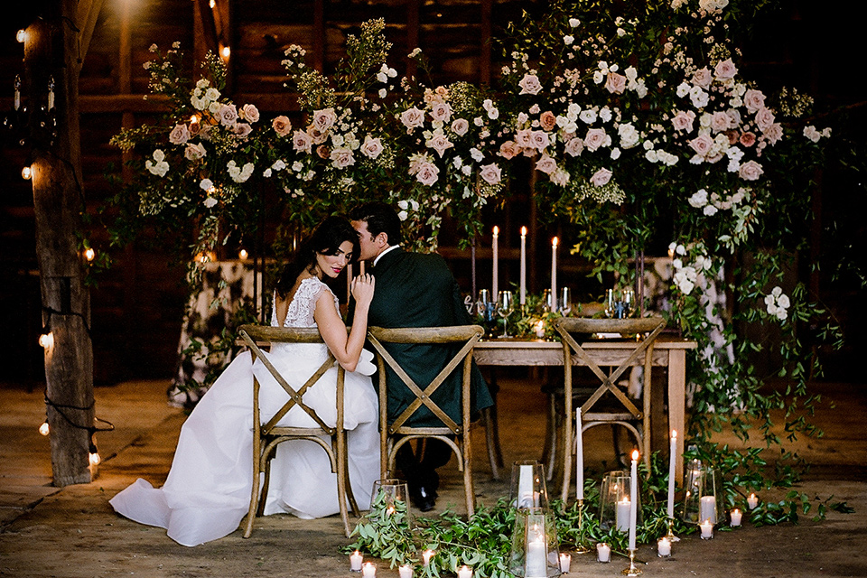 bride in a white ballgown with the groom in a green suit sitting at a farm table inside a barn
