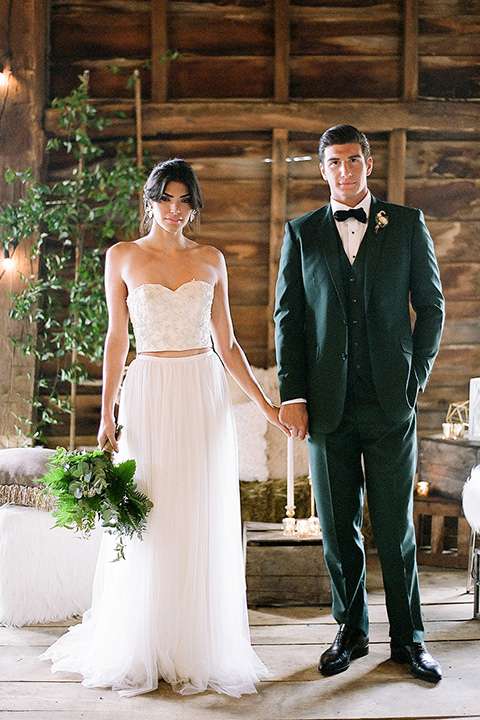  bride in a white two piece gown with a strapless neckline with her hair in a loose brain and garden bouquet the groom in a green suit walking in venue