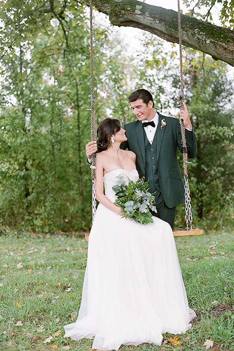  bride in a white ballgown with her hair in a loose brain and garden bouquet the groom in a green suit on the swing