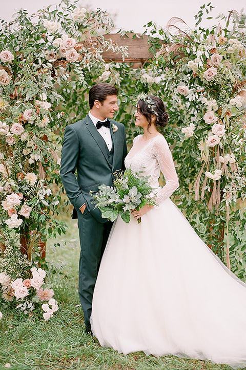  bride in a white ballgown with her hair in a loose brain and garden bouquet the groom in a green suit standing under the archway at the ceremony