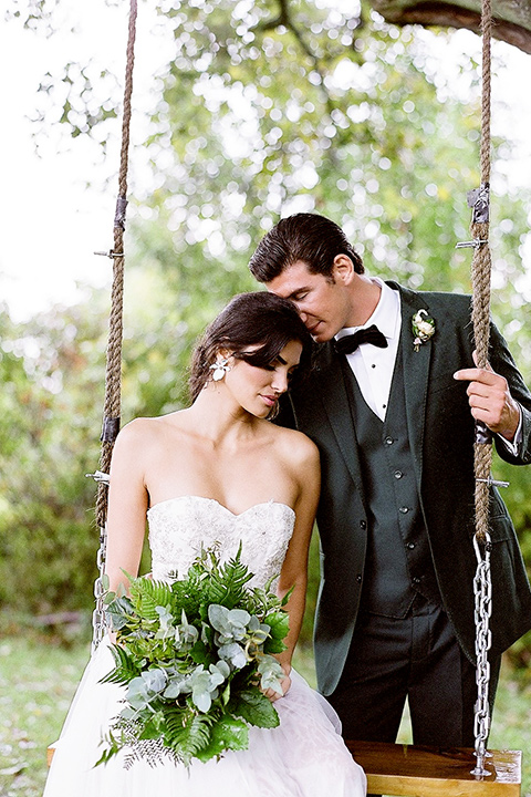  bride in a white ballgown with her hair in a loose brain and garden bouquet the groom in a green suit sitting on the swing together